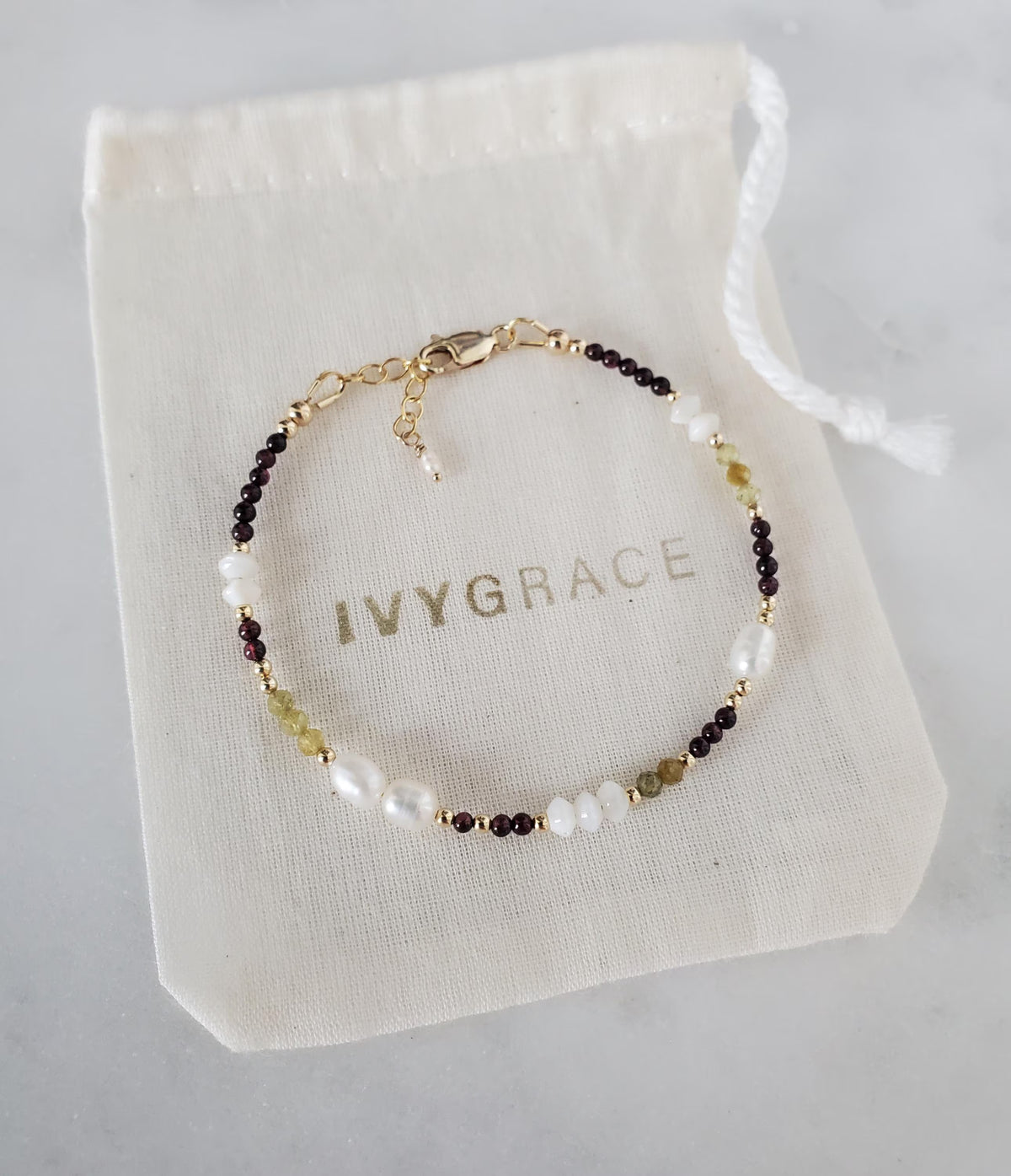 A delicate beaded bracelet with white pearls, maroon garnet, and green garnet beads with gold-filled detail.  Displayed on an ivory drawstring pouch with the brand name 'IVYGRACE' printed on it.